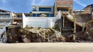 A coastal building with a staircase leading to a balcony, a sandy beach in the foreground, and other buildings in the background.