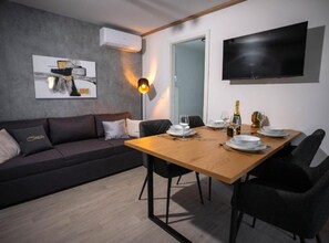 A modern living room with a dark sofa, a wooden dining table set for two, a mounted flat-screen TV, and an air conditioning unit on the wall. - Lux Mobile Homes Vodice (Vodice)