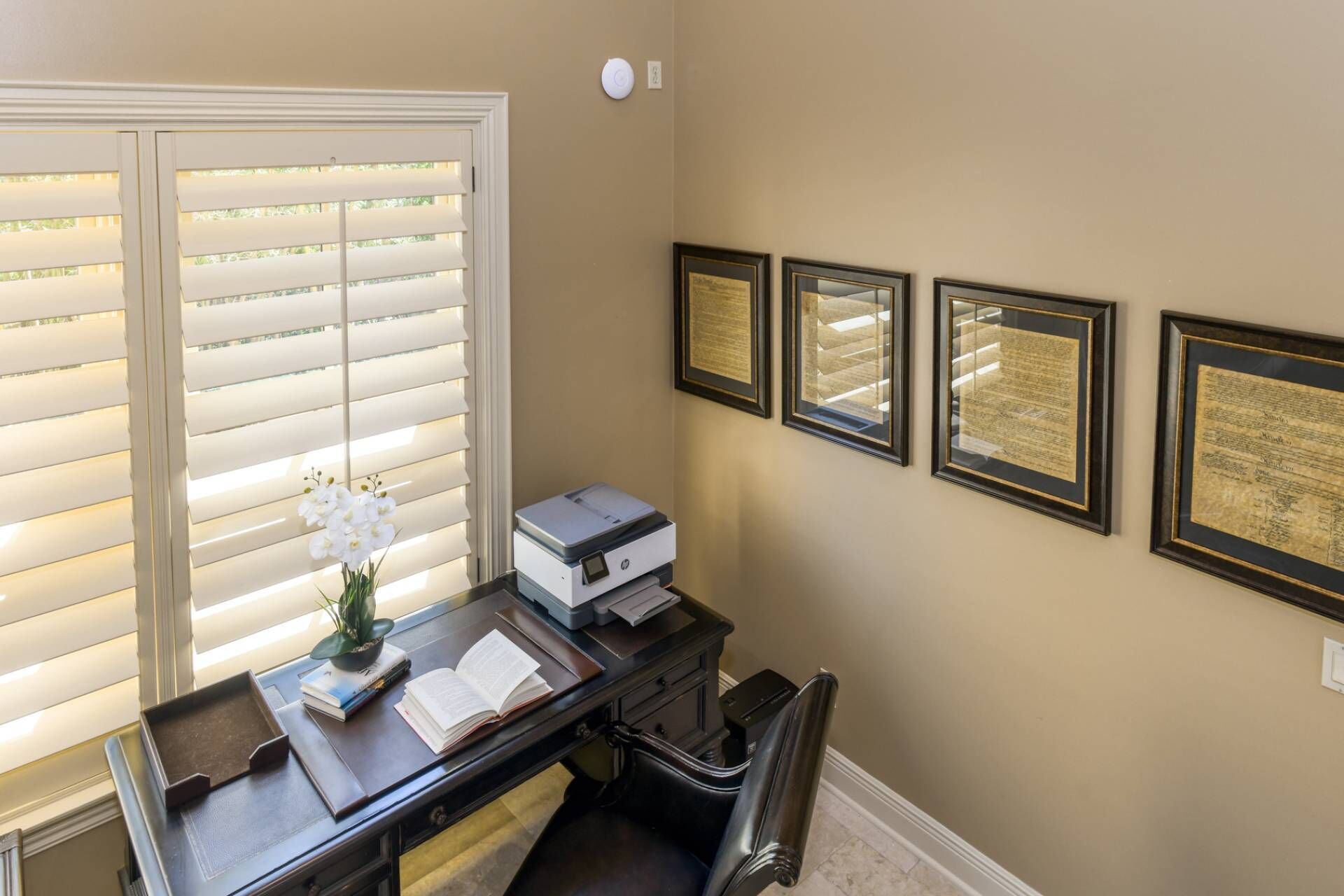 A room with a desk, a printer, a chair, and a window with blinds. There are four framed documents on the wall.