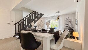 Ein Essbereich mit einem weißen Tisch, schwarz-weißen Stühlen, einer schwarzen Treppe und einem Fenster mit Blick auf Grün.