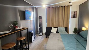 Een kleine kamer met een bed, een bureau met twee krukken, een televisie en een tafeltje met een plant.