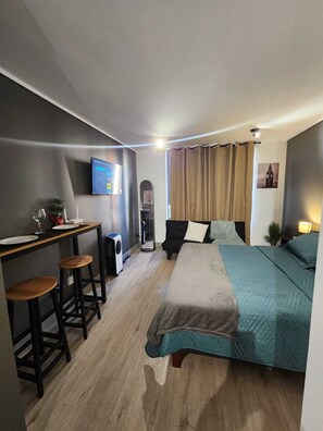Ein kleiner Raum mit einem Bett, einem Schreibtisch mit zwei Hocker, einem Fernseher und einem Tisch mit einer Pflanze.
