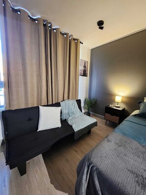 Ein Zimmer mit einer schwarzen Couch, einem Bett, einem Nachttisch mit Lampe und einer Wandleuchte.