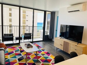 Ein modernes Hotelzimmer mit großem Fernseher, einem Balkon mit Meerblick und einem farbenfrohen, geometrischen Teppich.