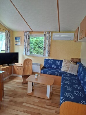 A compact living space with a sofa, TV, wooden table, and a window with curtains. - Lovely mobile home carcans in 3-star campsite (Carcans)