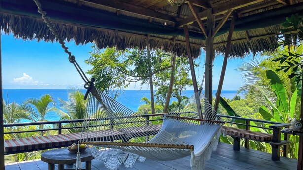 Un hamac sur une terrasse en bois, avec vue sur l’océan et une végétation luxuriante.