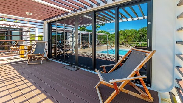 En udendørs terrasse med en glasdør, der fører til et poolområde, træterrasse og to liggestole.