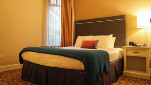 Una habitación de hotel con cama, mesita de noche, lámpara y ventana con cortinas.