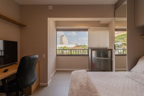 Quarto com cama, escrivaninha com televisão e varanda com vista para prédios.