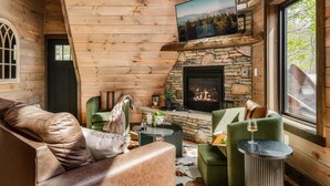 Et hyggeligt interiør i en træhytte med en pejs af natursten, en lædersofa og et fjernsyn monteret over sofaen.