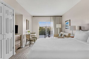 Habitación de hotel con cama, escritorio, silla, televisor, balcón con cortinas y ventana con vistas.