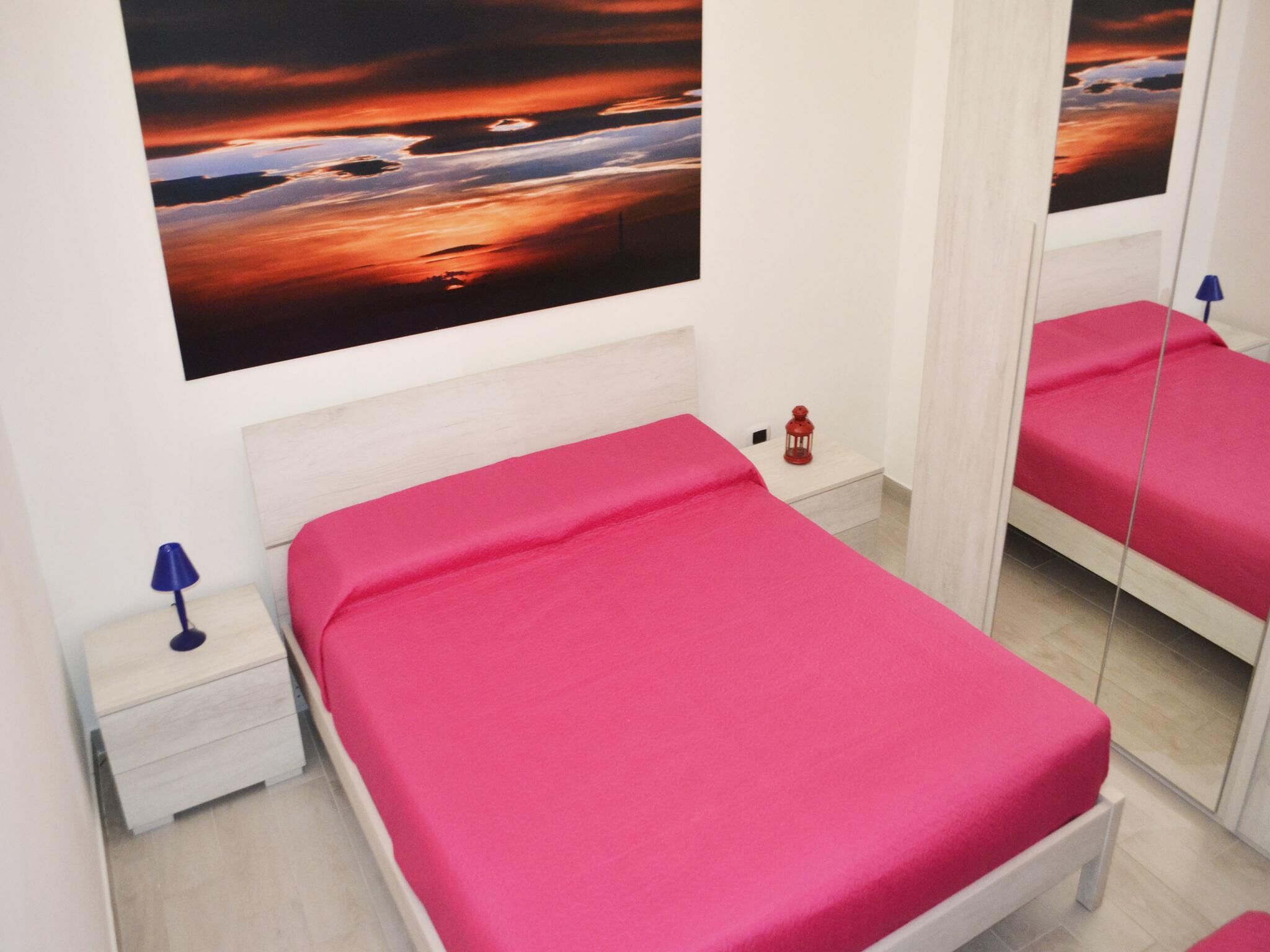 Une chambre à coucher avec un lit, une table de chevet, une lampe, un miroir et un tableau représentant un coucher de soleil accroché au mur.