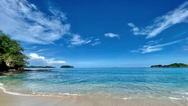 Ein Strand mit klarem Wasser, entfernten Inseln und blauem Himmel.