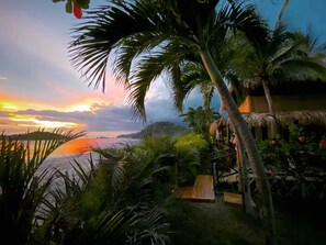 Ein tropisches Resort mit Holzterrasse, Palmen und Blick auf den Ozean bei Sonnenuntergang.