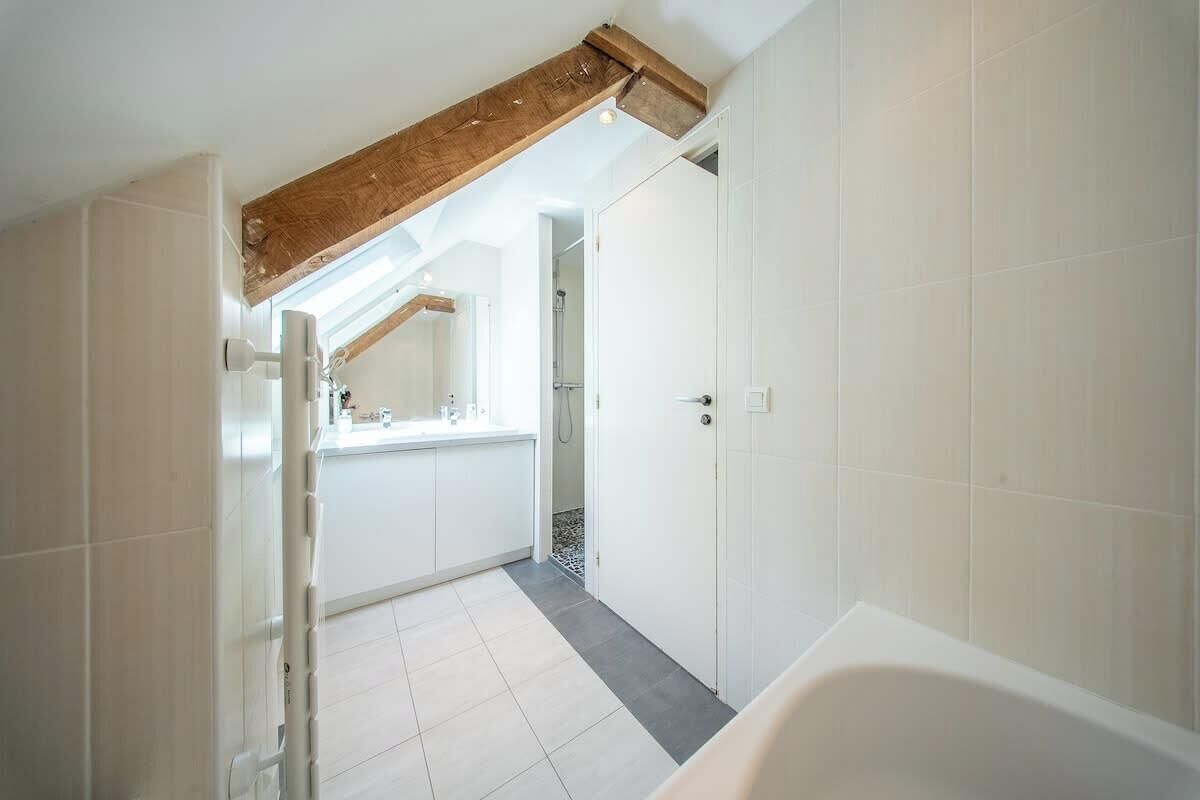 A bathroom with a bathtub, white tiles, a wooden beam ceiling, and a door leading to another room.