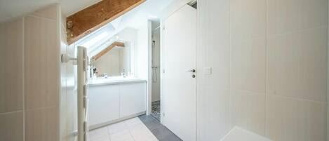 A bathroom with a bathtub, white tiles, a wooden beam ceiling, and a door leading to another room.