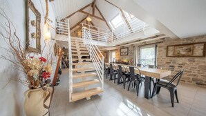 A spacious loft with a high ceiling, exposed wooden beams, a central staircase, a dining area with modern chairs, and a stone wall. - Grande longère entre campagne et océan (Guidel)