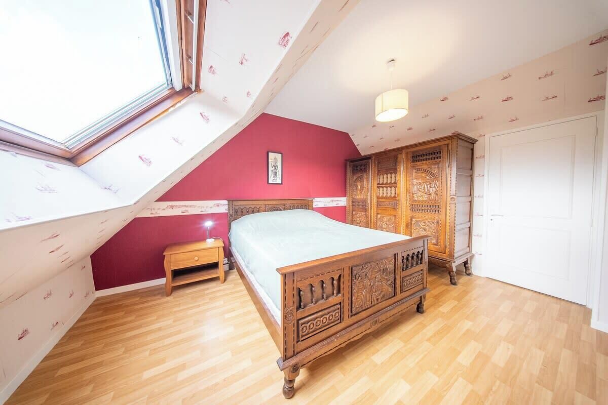 Ein Schlafzimmer mit schrägen Wänden, einem hölzernen Bett, einem Nachttisch, einem Kleiderschrank und einem großen Fenster.
