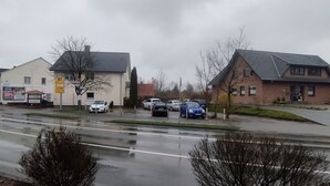 Una veduta di strada con una fila di auto parcheggiate, un edificio con un cartello pubblicitario e una strada bagnata, segno di pioggia recente.