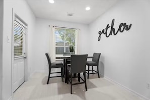A dining area with a table and chairs, a window with blinds, and a wall with the word 'gather' written on it.
