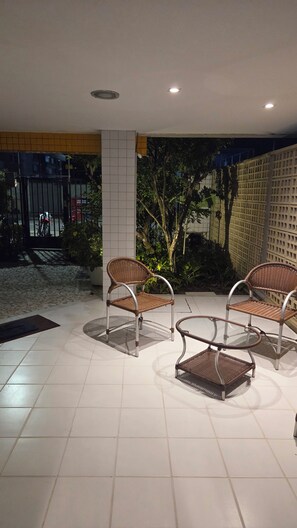 A covered outdoor seating area with wicker chairs and a glass table, surrounded by tiled flooring and potted plants. - Cabo Branco apartment in João Pessoa, 180 meters from the beach. (JOAO PESSOA)