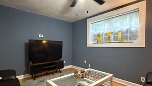 A living room with a ceiling fan, a television, a coffee table with decorative items, and a window with letters on the wall. - West Midtown Bungalow Pup Friendly (Atlanta)