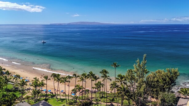 A beach with clear blue water, palm trees, and a boat in the distance. - Beautiful remodeled Kihei Condo next to the Beach! (Kihei)