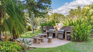 An outdoor patio with a grill, stone tables, and lush greenery. - Beautiful remodeled Kihei Condo next to the Beach! (Kihei)