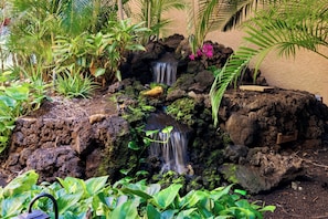 Ein Garten mit einem kleinen Wasserfall, umgeben von verschiedenen Pflanzen und Steinen.