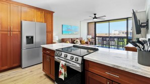 A modern kitchen with wooden cabinets, a stainless steel refrigerator, a gas stove, and a large window overlooking a balcony. - Beautiful remodeled Kihei Condo next to the Beach! (Kihei)