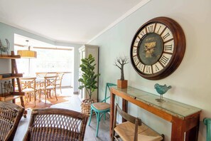 A room with wicker furniture, a wooden table, a large wall clock, and a view through a window.