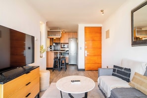A modern living room with a flat-screen TV, a sofa, a coffee table, and a kitchen in the background. - HOM | Metro | Las Condes | Calefacción | Parking (Las Condes)
