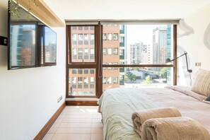 A bedroom with a bed, a TV, a window with a view of buildings, and a lamp. - HOM | LasCondes | Metro Manquehue | Parking | 1D1B (Las Condes)