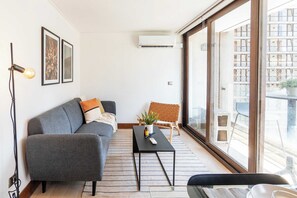 A modern living room with a grey sofa, a small black table, a potted plant, and a large window. - HOM | LasCondes | Metro Manquehue | Parking | 1D1B (Las Condes)