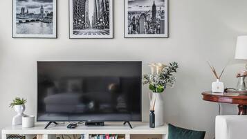 A living room with a flat-screen TV, framed cityscape pictures, a bookshelf, and a round table with a vase.