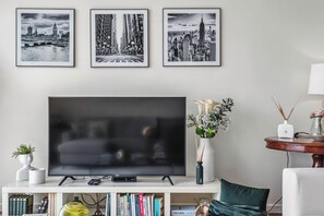 A living room with a flat-screen TV, framed cityscape pictures, a bookshelf, and a round table with a vase.
