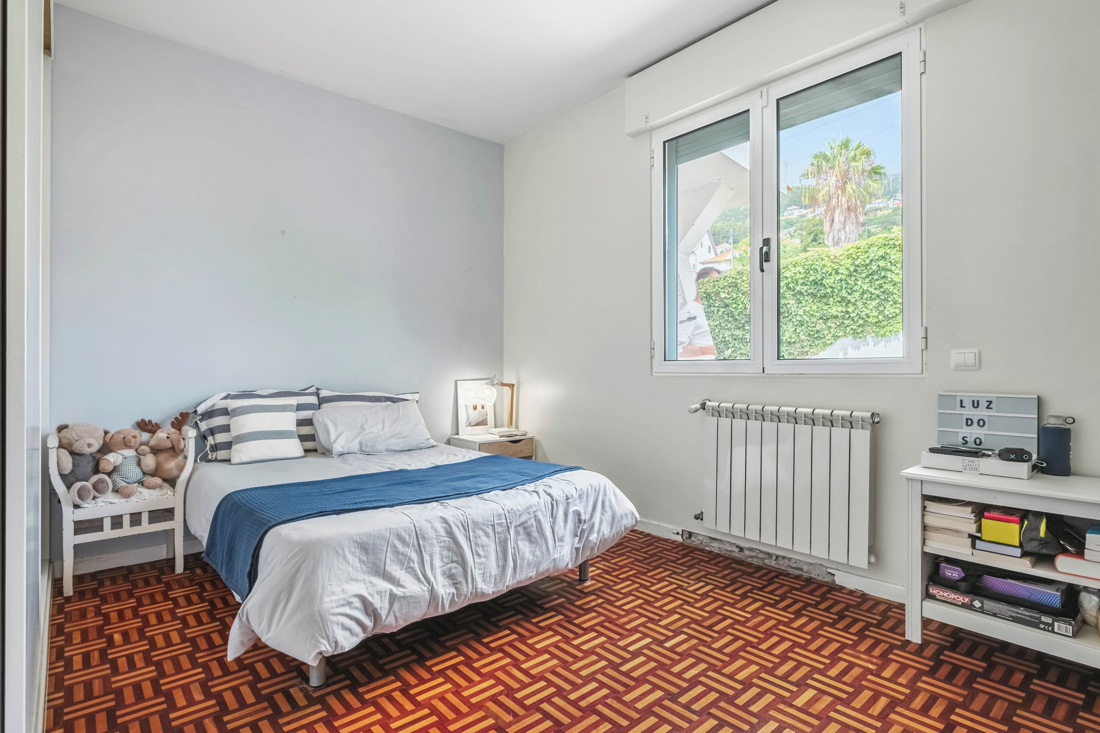 A bedroom with a bed, a nightstand, a window with a view, a radiator, and a shelf with books.