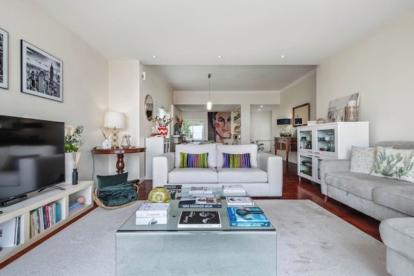 Ein modernes Wohnzimmer mit Fernseher, einem Couchtisch mit Büchern, einem weißen Sofa und einem Essbereich im Hintergrund.