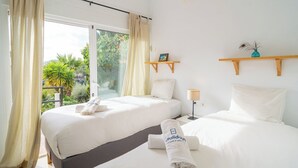 Habitación de hotel con dos camas, una ventana con cortinas, una mesita con lámpara y vistas al exterior con vegetación.