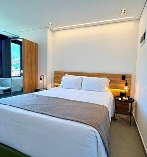 A modern hotel room with a large bed, a wooden headboard, a bedside table with a lamp, and a view of the city through a large window.