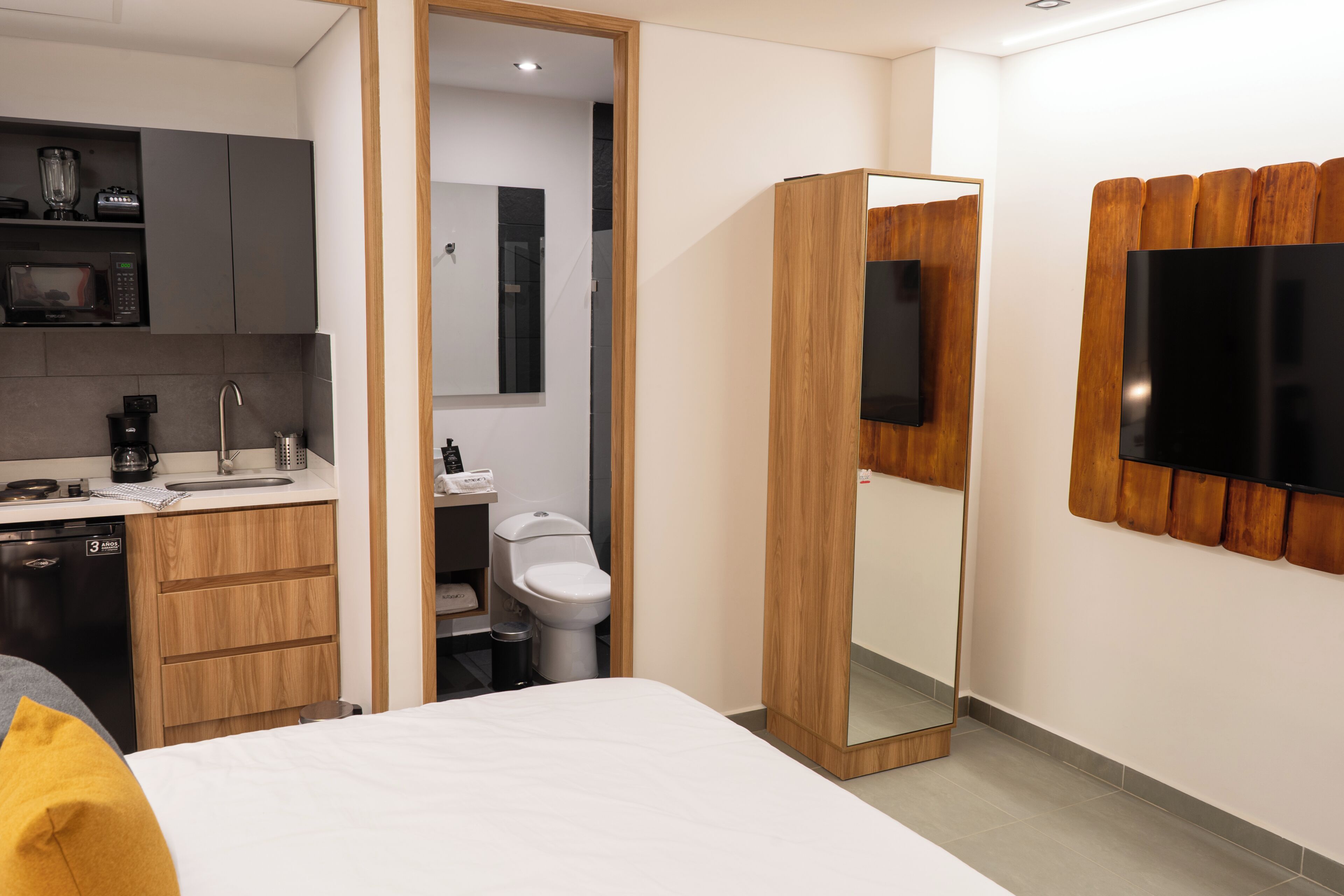 A modern hotel room with a bed, a wooden wardrobe, a built-in TV, a kitchen area with a microwave and coffee maker, and a bathroom with a toilet and sink.