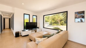Ein modernes Wohnzimmer mit einem großen Fenster, einem Flachbildfernseher, einem beige gefärbten Sofa und einer weißen Unterhaltungsanlage.