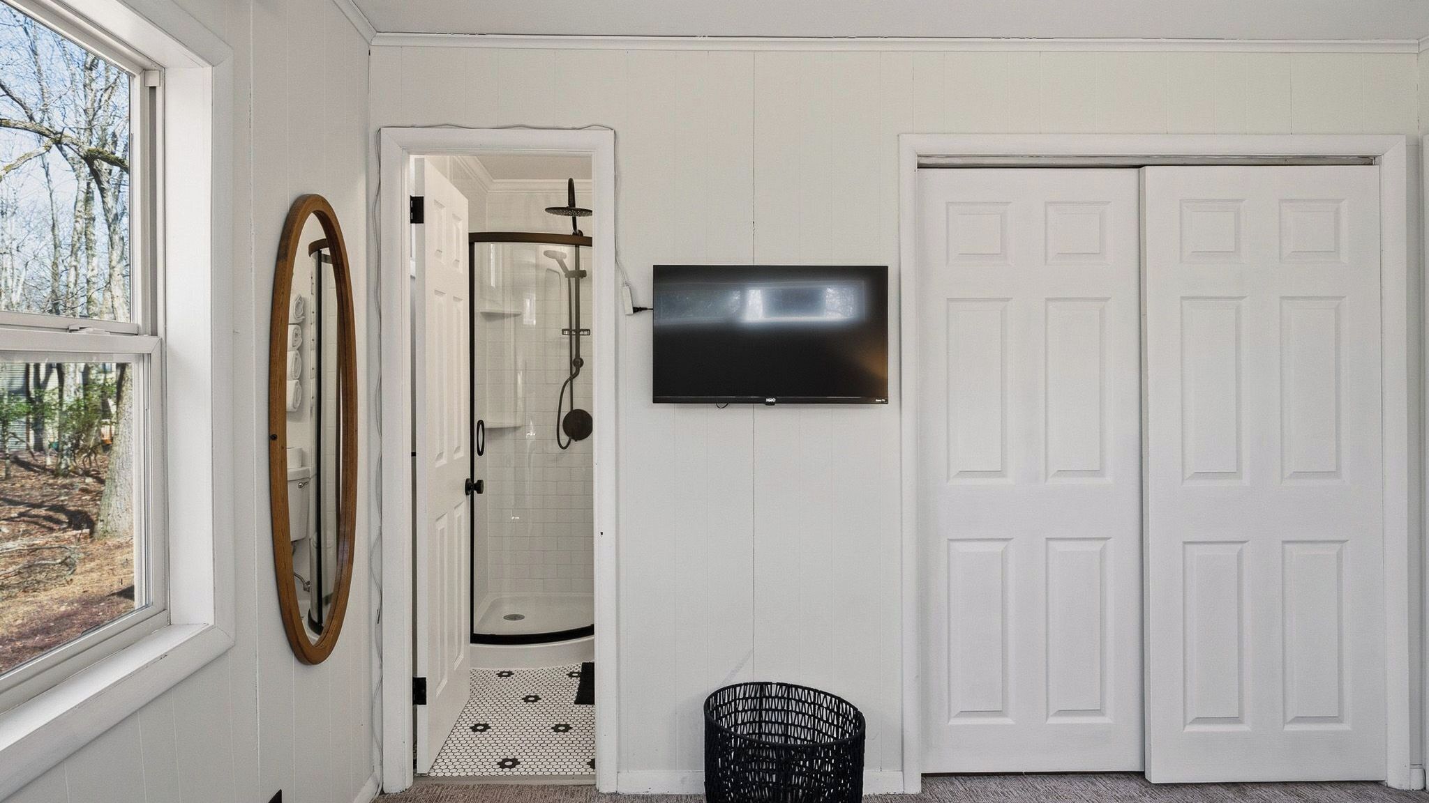A bathroom with a shower, a toilet, a sink, a mirror, a television, and a door.