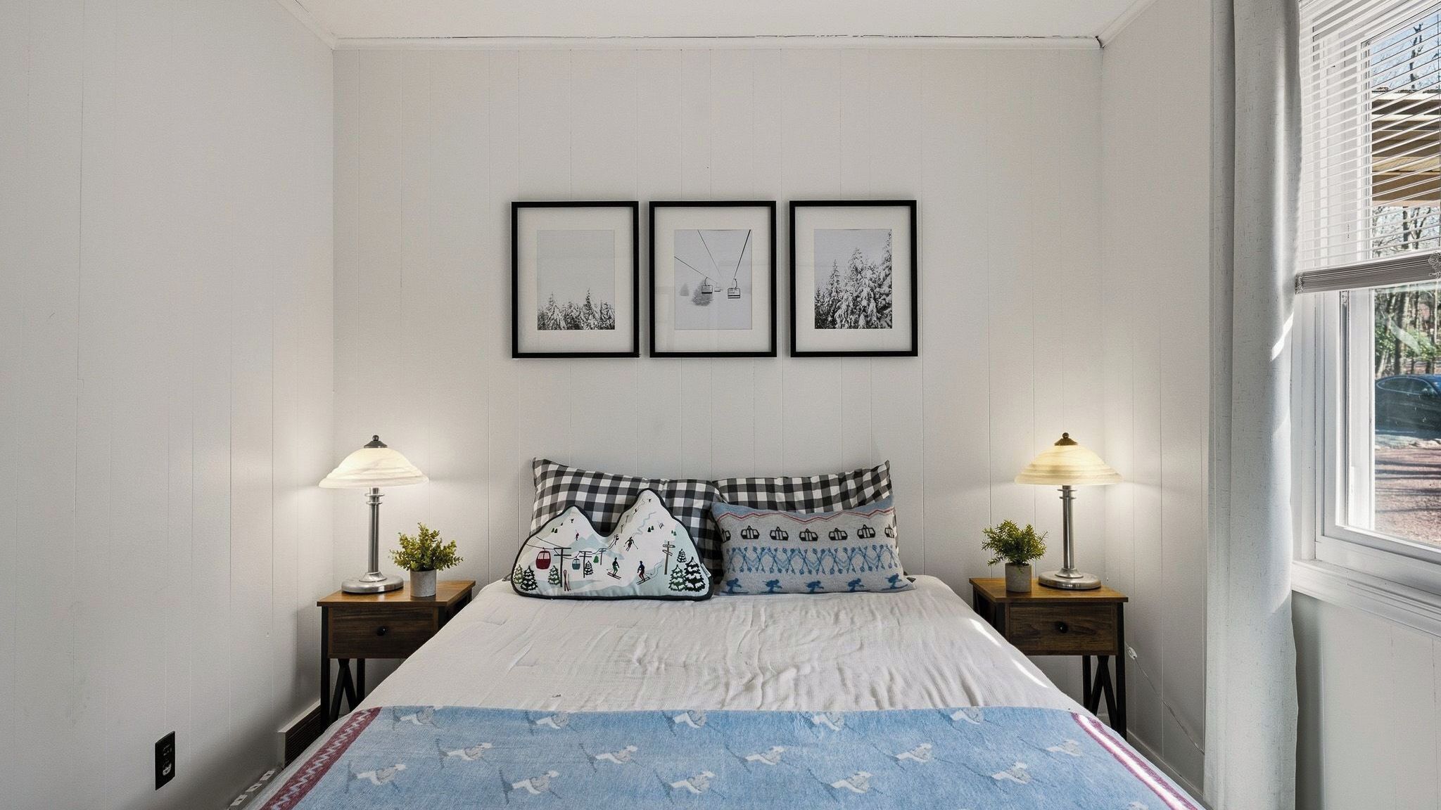A bedroom with a bed, two bedside tables with lamps, a window with blinds, and three framed pictures on the wall.