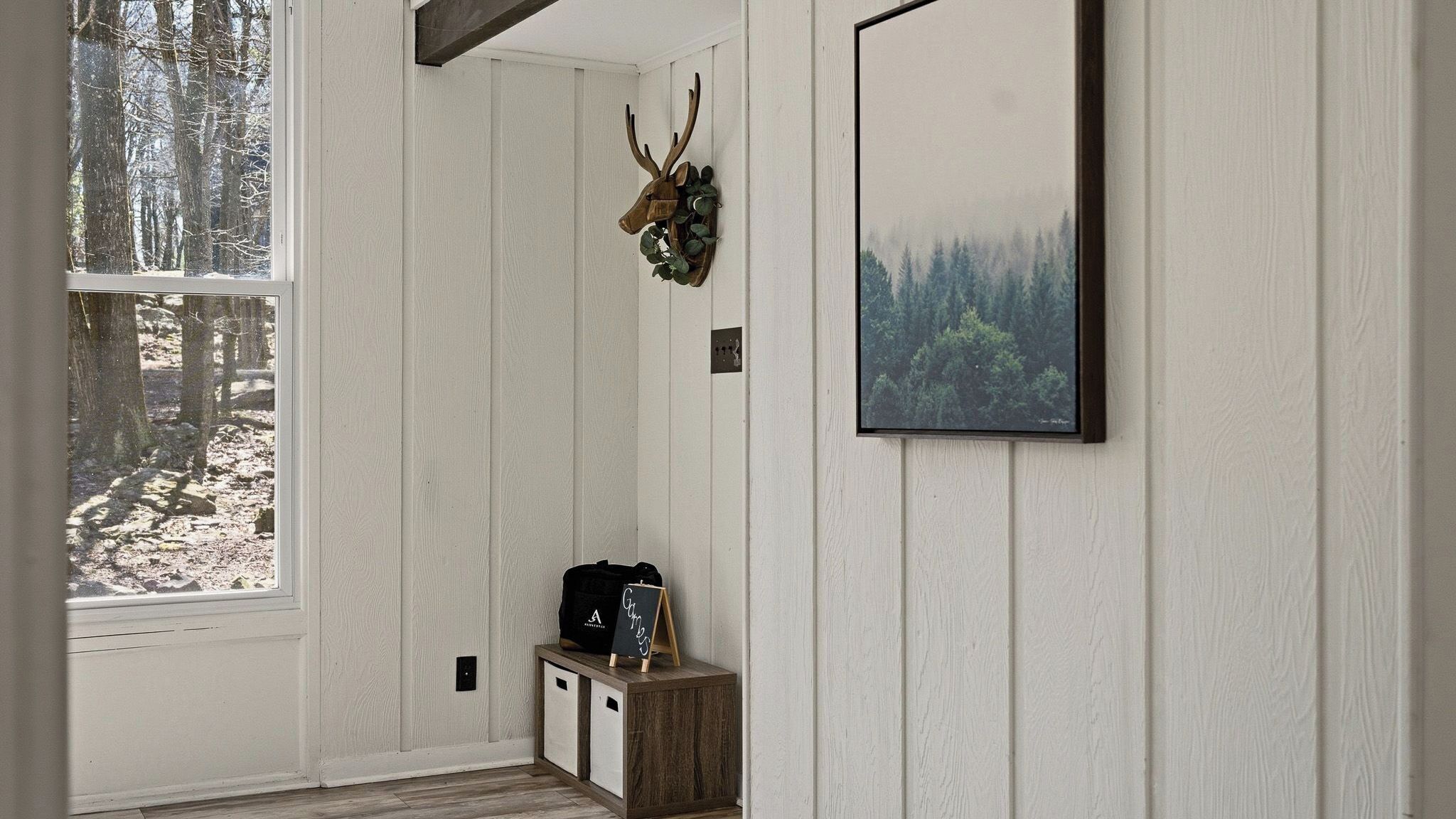 A room with a window overlooking a forest, a mounted deer head, a framed forest landscape, and a small wooden cabinet with a black object on top.