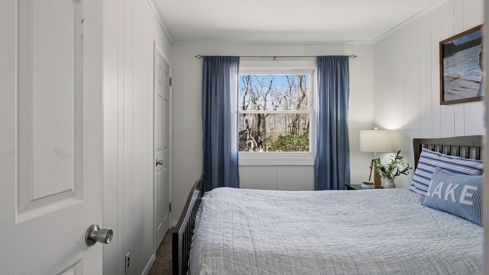 Een slaapkamer met een bed, een raam met gordijnen, een nachtkastje met een lamp en een aan de muur hangend schilderij.