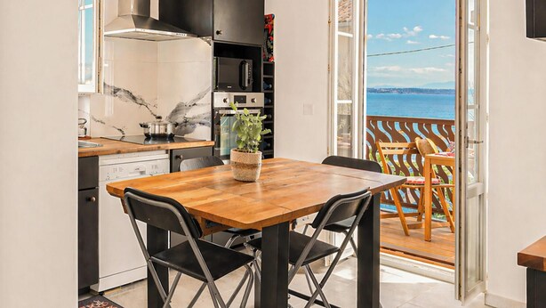 A modern kitchen with a wooden dining table and chairs, a view of the sea through an open door, and a balcony with outdoor seating. - Casa Turturella – Vue mer et citadelle au cœur de Calvi (Calvi)