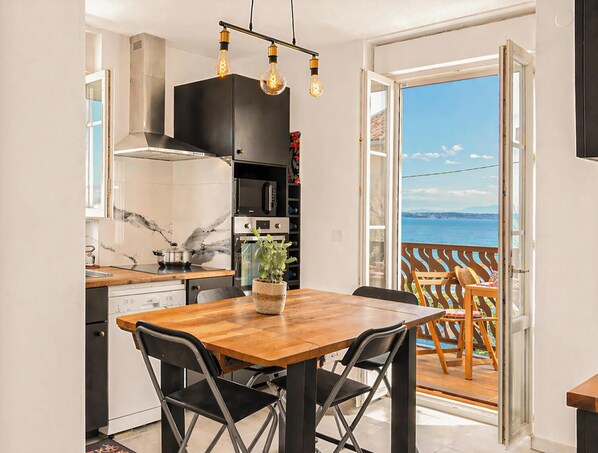 Ett modernt kök med ett matbord och stolar i trä, utsikt över havet genom en öppen dörr och en balkong med utemöbler.
