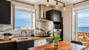 A modern kitchen with a large window, a wooden dining table, and a view of the sea. - Casa Turturella – Vue mer et citadelle au cœur de Calvi (Calvi)