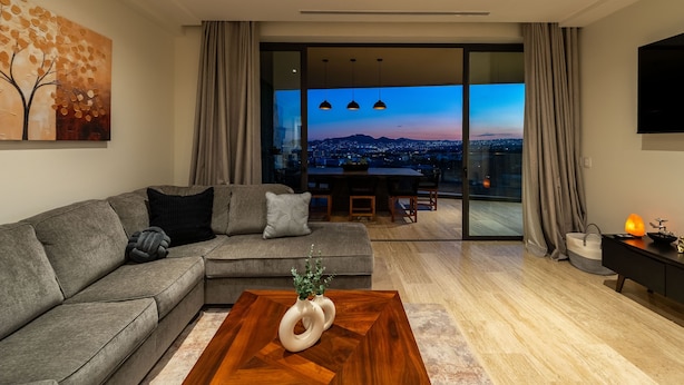 A modern living room with a sofa, a coffee table, a painting, a dining area, and a view of the city at dusk. - Mistiq Views in Cabo San Lucas (Cabo San Lucas)